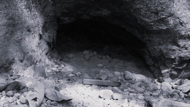 Monochrome image of a dark natural cave beneath a rocky overhang, surrounded by rounded stones, fallen leaves, and rugged geological surfaces.