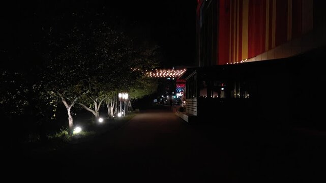 A dark pedestrian path runs past illuminated trees and a modern building terrace at night. 