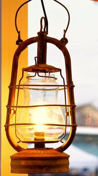Old rusty kerosene lantern burning bright near window glass frame surface view. Retro metal oil lamp emitting warm golden fire light illumination inside rustic country house room habitat.