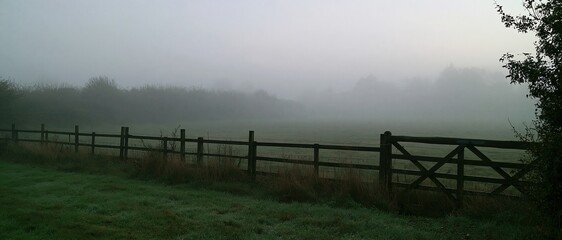 Naklejka premium Autumn fog covering a grey field with a damp timber fence line and a dim hedge, a muted atmospheric background for compositing and copy space