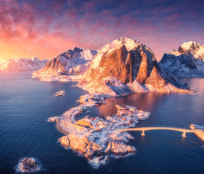 Aerial drone view of Hamnoy fishing village in Lofoten, Norway. Winter arctic landscape with snow covered mountains, bridge, islands, ocean sea, pink sky, clouds and red sunset sunlight. Top view