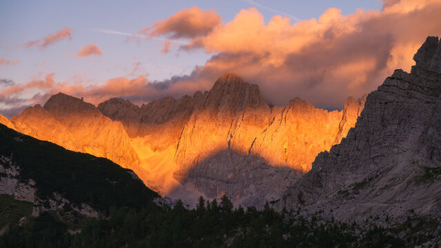 Golden sunset light on rocky mountain peaks in Slovenia in autumn. Alpine landscape of Julian Alps with pink sky, orange clouds, sunbeams, steep cliffs, trees slope and dramatic evening sunlight