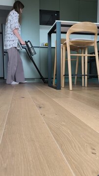 Stick vacuum cleaner with blue led light used by woman to clean engineered wood flooring. Female housewife vacuuming parquet floor surface in modern kitchen living room interior. Everyday