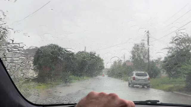 POV of male hand on steering wheel driving a car during a heavy rain on rural road. Detailed view from inside the vehicle with active windshield wipers clearing wet glass on spring rainy day. Slow mot