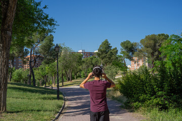 Photograph from a rear perspective of a young man walking along a park path while listening to music with headphones. © Laura