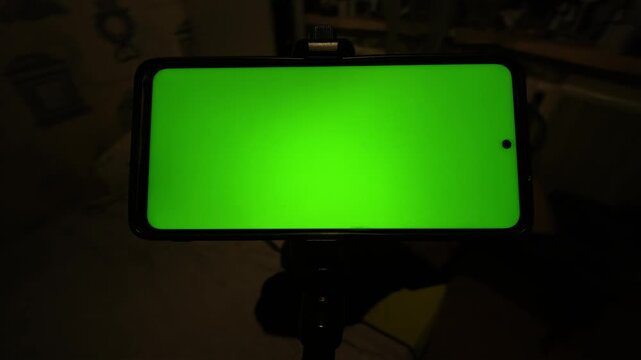 A smartphone with a bright green screen placeholder is mounted horizontally on a tripod in a dark room. 