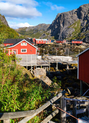 Traditional red fishing houses of Nusfjord stand by calm water, surrounded by steep rocky mountains and autumn nature. © EKH-Pictures