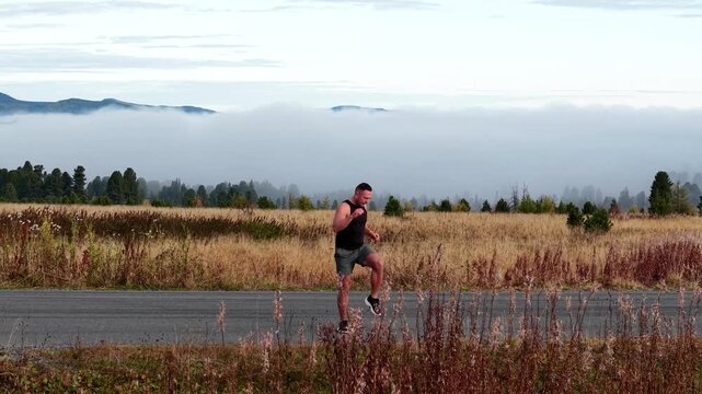 Drone cinematic parallel tracking shot of an athletic man performing high-knee warming up exercise on a mountain road.