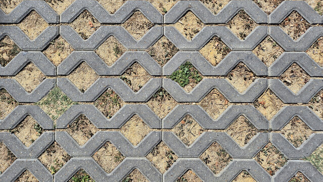 A top-down view of grey interlocking concrete grid pavers with open diamond-shaped holes filled with dirt and sparse green grass patches.