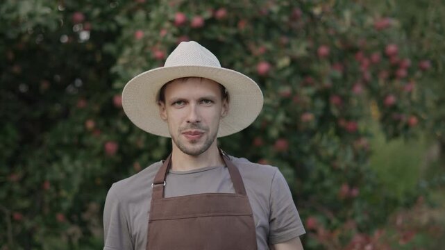 Caucasian man in hat in apple orchard has fun on camera with apples in hands. Portrait of farmer in good mood fools around during harvest.