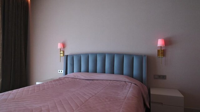 Serene bedroom corner features soft pink quilted bedspread, blue tufted headboard, minimalist white nightstand, and gold sconces with pink shades