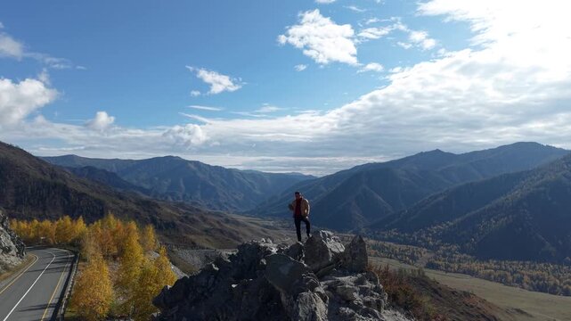 Drone panning from right to left tracking a man standing on a high rocky peak overlooking a scenic mountain valley.
