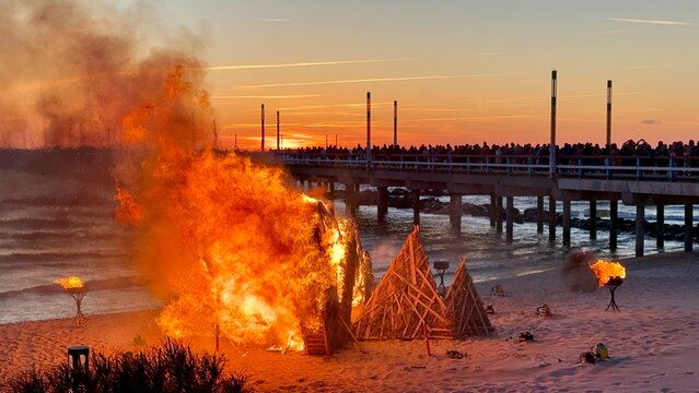 Dramatic scene of Jurginės celebration in Palanga, Lithuania, featuring burning straw sculptures on the beach near the sea pier during a vivid sunset.