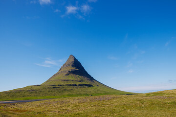 Fototapeta premium Iconic cone-shaped mountain Kirkjufell, Snæfellsnesvegur, Iceland rising from green grassland under a clear blue sky, with a winding road and soft white clouds in Iceland.