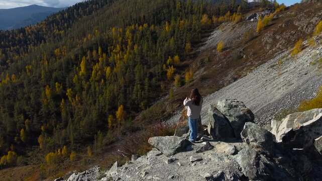Drone panning from right to left tracking a woman standing on a rocky peak and looking at an autumn forest.