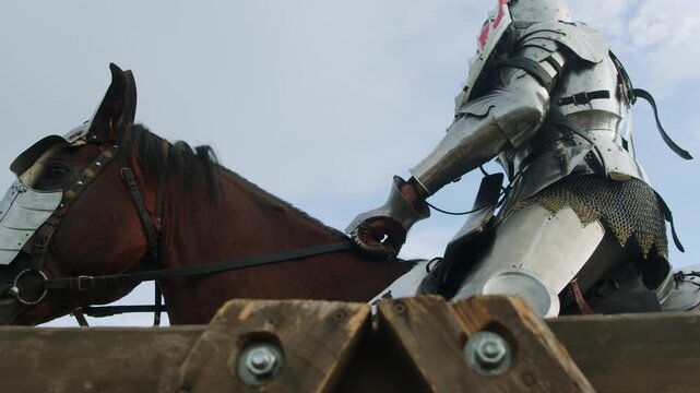 Armored knight on horseback rides slowly along wooden fence. Low angle shot captures shining metal gear and decorated shield against cloudy blue sky background during medieval historical festival.