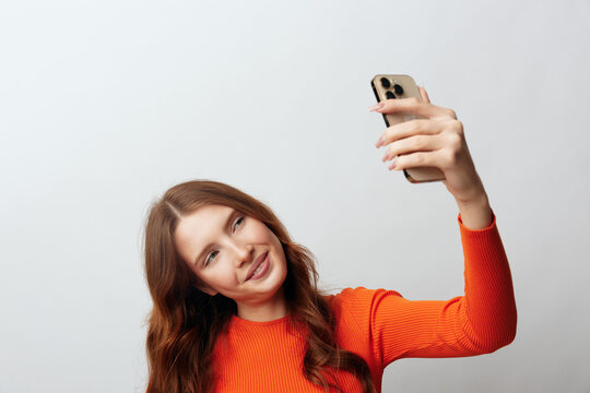 Young woman with long red hair in an orange sweater takes a selfie with her smartphone, smiling happily. Studio shot on a gray background. Concept of social media and self expression.