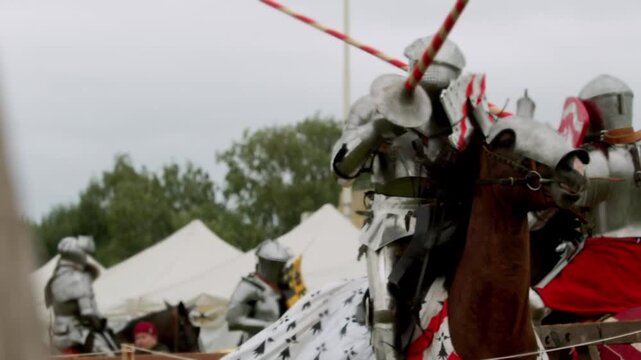 Splintering wood flies as heavy cavalry crashes on tournament field. Opposing competitors riding strong brown horses smash long poles against metal armor and red shield.