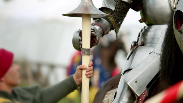 Assistant attaches red painted shield onto mounted knight. Shiny steel armor protects medieval warrior preparing for jousting tournament near historical white tower.
