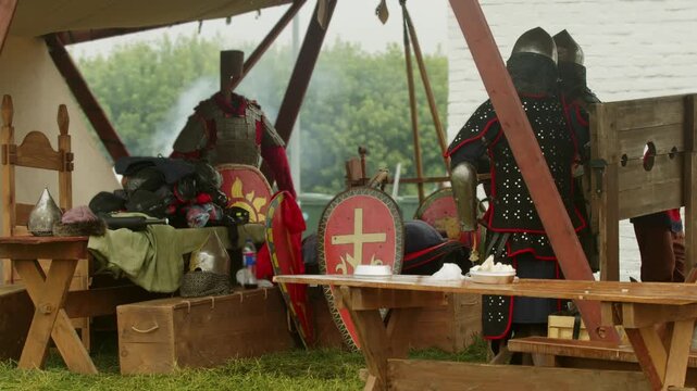 Authentic medieval armor stands under canvas tent roof. Red shields and heavy metal helmets rest on wooden tables during historical festival reenactment event.