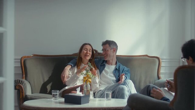 Reconciled couple smiling happily, embracing and talking to psychologist after resolving their conflict during successful family therapy session