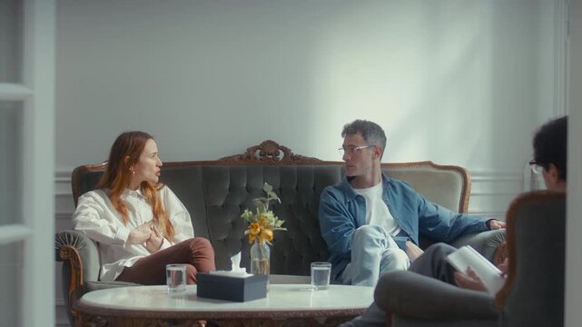 Married couple sitting on sofa, arguing emotionally and blaming each other during relationship therapy session with psychologist