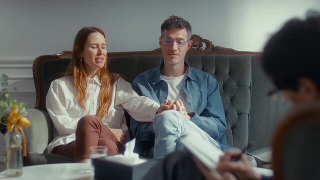 Happy married couple sitting close together on sofa, smiling and holding hands as speaking about positive breakthrough with therapist during successful counseling session