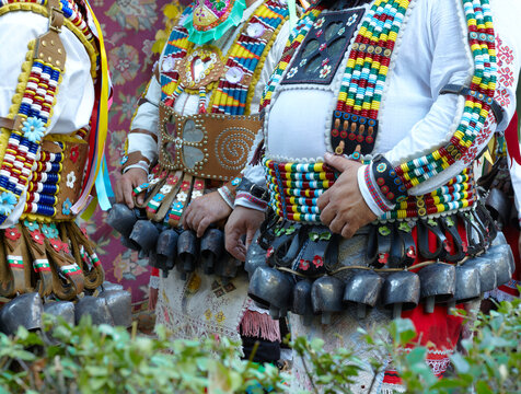 Bulgarian kukeri dancers in colorful beaded costumes with large metal bells, leather belts, and layered traditional garments at an outdoor cultural gathering