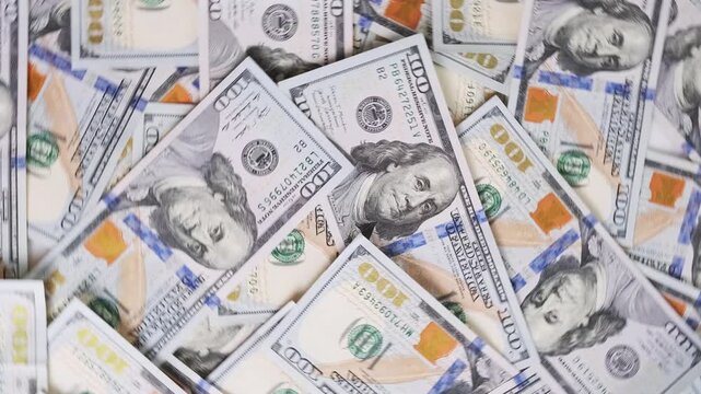 Rotating United States hundred dollar bills create dense money background, with overlapping banknotes scattered across frame, changing orientation in continuous pattern of cash and currency