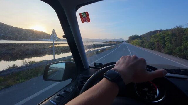 POV drive with one hand on the steering wheel along a country road past the flooded salt pans of Ses Salines as the low sun sets behind pine covered hills, Ibiza, Balearic Islands, Spain