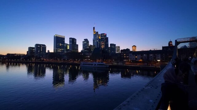 Frankfurt skyline at blue hour with illuminated skyscrapers reflected in the calm river Main, an excursion boat at the quay, seen from the Eiserner Steg footbridge, Frankfurt am Main, Hesse, Germany