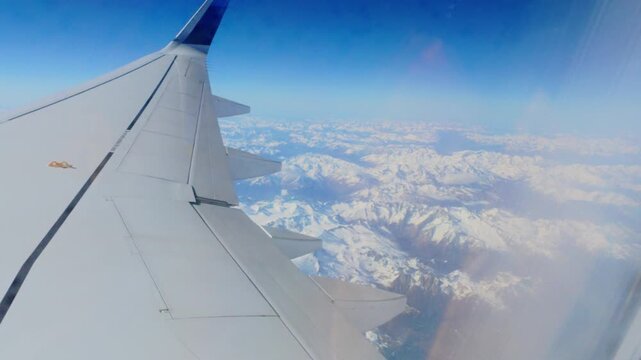 Airplane wing in cruise flight above snow covered Alps mountain ranges and valleys seen through the cabin window under a deep blue sky, Alps, Europe