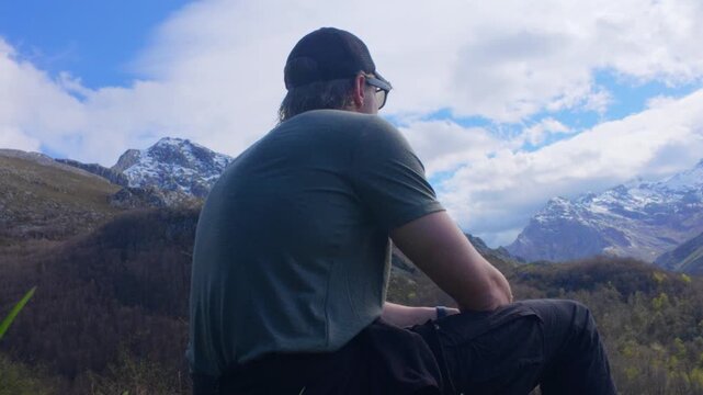Man in a cap and t shirt seen from behind sitting on a grassy slope and gazing across a wooded valley to snow capped peaks under drifting clouds, low locked off shot, Picos de Europa, Spain