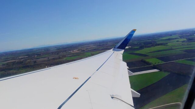 Airplane climbing after takeoff seen through the cabin window, wing over green patchwork farmland and roads with the Alps faintly visible on the hazy horizon, near Munich, Bavaria, Germany