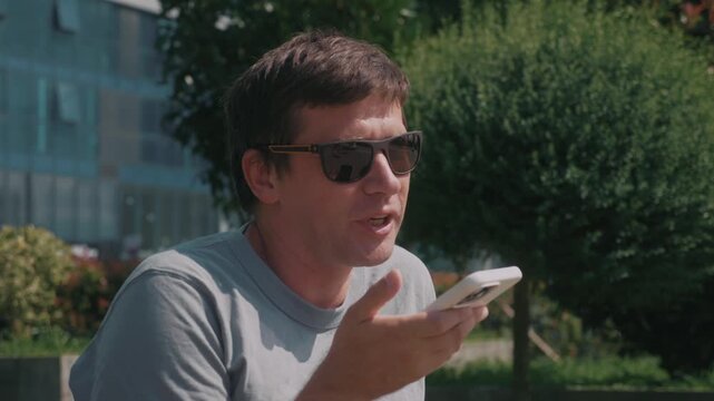 Man records voice message on smartphone for mobile communication, speaking into phone and sending spoken update through messaging app outdoors.