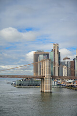 Fototapeta premium A tower of Brooklyn Bridge rises beside the Manhattan waterfront. Stone arches overlook gray river water with a glass pier building below a clouded blue sky.