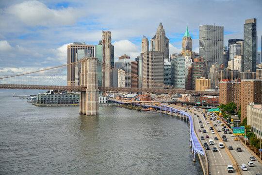 Brooklyn Bridge crosses the river beside a highway below Manhattan towers. Cars follow a curving shoreline highway below towers as blue gaps appear between gray clouds.