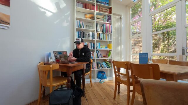 Static wide shot of a young man in a cap working on a laptop at a wooden table in a bright library cafe with bookshelves and garden windows, Frankfurt am Main, Hesse, Germany