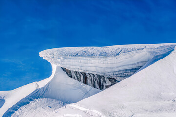 A huge wind sculpted snow cornice curls over a dark ice streaked rock face beneath a deep blue mountain sky © Kafi