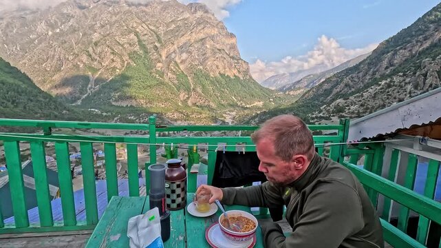 Man enjoys breakfast on green balcony overlooking mountain valley. He drinks coffee while staring at scenic view. Peaceful morning moment with natural beauty. Ideal for travel, adventure