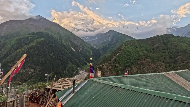 Nepal flag flutters against Annapurna peak at dusk. Green rooftops frame mountain valley below. Distant peaks glow with golden sunset light. Flags add cultural touch to high-altitude scene