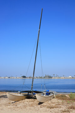 Small catamaran sailboat with a tall black mast sits on the sandy shore of Mission Bay in San Diego, California