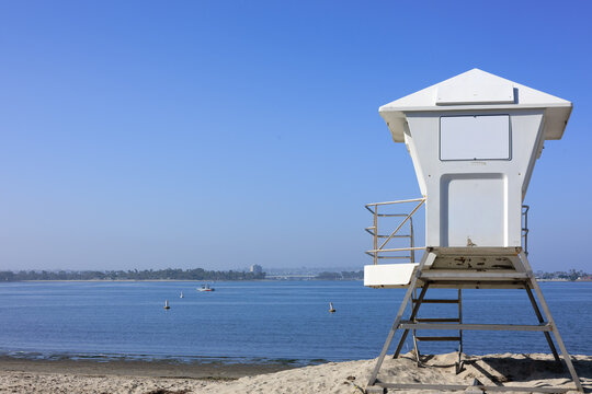 A white painted lifeguard tower sits on a sandy shoreline overlooking the calm blue waters and distant houses of Mission Bay in San Diego, California.