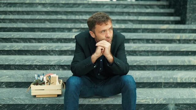 Distressed male worker facing lost work sitting quietly on outdoor stairs beside box. Poverty risk and layoff