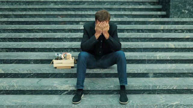 Laid off male worker after downsizing hiding face on building stairs outdoors. Burnout and depression
