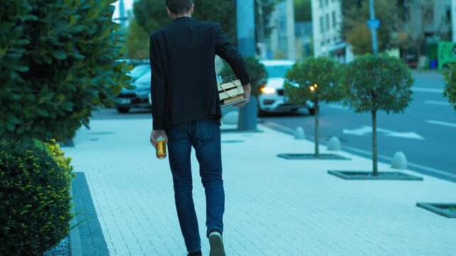 Redundant male worker walking on street drinking beer carrying office box. Career collapse and poverty