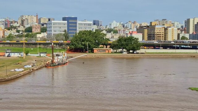 Water dredging vessel operating near a lush riverbank in Porto Alegre Brazil with a wide urban backdrop of highways, overpasses, and city buildings under a blue sky