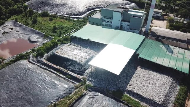 Aerial drone wide shot of a waste management and recycling facility surrounded by lush green mountains and forest. The footage shows industrial buildings and waste processing areas within a rural land