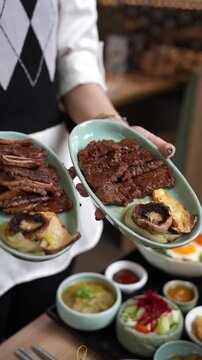 Close-up 4K footage of waitress hands holding two plates of Korean BBQ galbi kalbi grilled beef short ribs with mushroom onion and banchan side dishes on restaurant table.