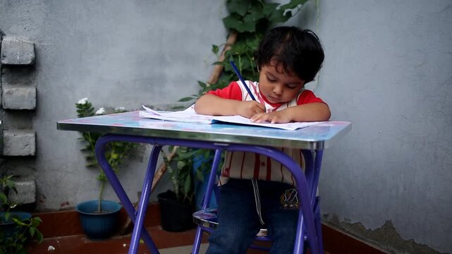 Elementary age cute child of Indian ethnicity sitting on chair near brick wall at home and studying book and writing alphabets in notebook video portrait close up.